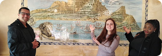 Three students smiling and gesturing towards a tiled coastal landscape mural