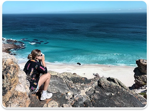 Student sitting on a rocky cliff photographing a turquoise beach and ocean below